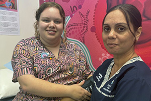 Aboriginal Health Practitioner Kira Prosser sits with an Aboriginal women in front of colourful Aboriginal artwork.