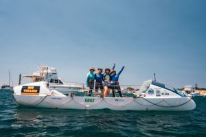 Landscape picture of the row boat taken lengthways in the ocean, with the four women standing together in the middle of the boat smiling to the camera. 