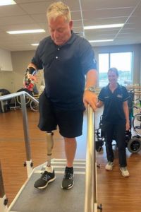 Dean walking up and down some stairs with his prosthetic leg in the SRS gym, with a physio in the background guiding him.