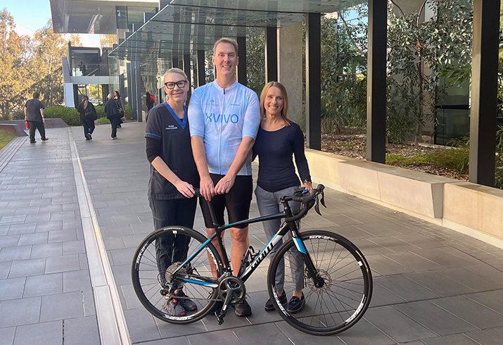 Heart transplant recipient Brett Driessen stands behind a bicycle. A nurse stands to his right and FSH Cardiac Transplant Nurse Practitioner Clare Fazackerley stands at his left