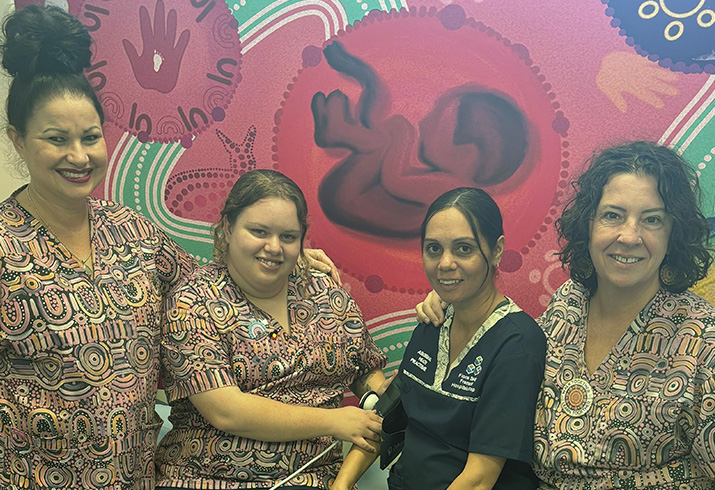 Aboriginal Health Practitioner Kira Prosser (centre left) sits with an Aboriginal women (centre right) in front of colourful Aboriginal artwork. Two other Boodjari Bidi clinic midwives stand with them. Kira and the clinic midwives wear scrubs with colourful Aboriginal designs