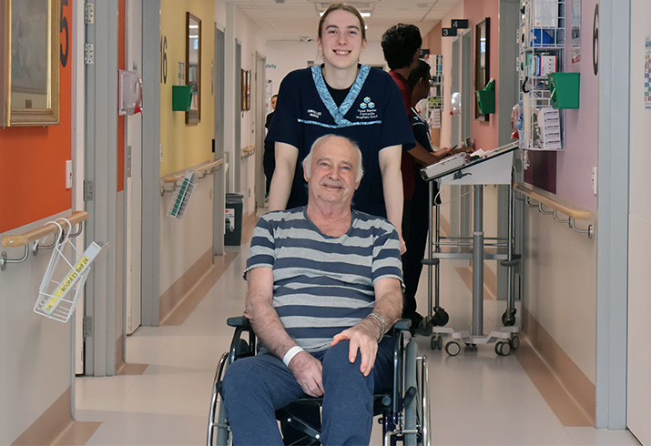 A male enrolled nurse pushes an older man in a wheelchair down a hospital corridor