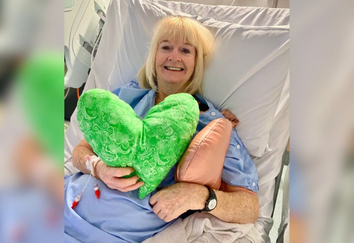 Keryl laying in a hospital bed wearing a blue patient gown, holding a green love heart shaped pillow. 