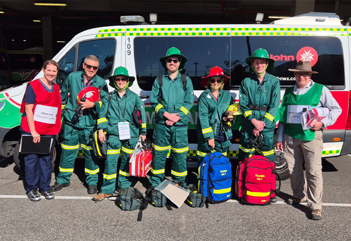 Seven staff from Fiona Stanley Hospital standing in front of an ambulance wearing green overalls and hardhats.
