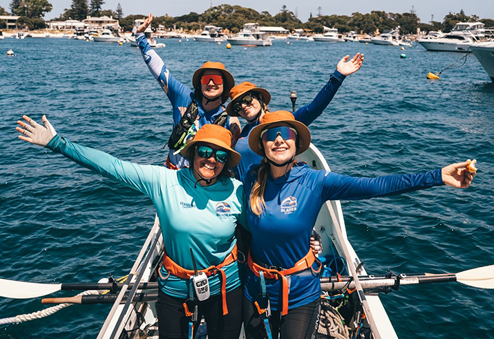 Four women standing on a rowing boat off the coast of Rottnest Island, wearing long sleeves, hats and sunglasses after a training session. 