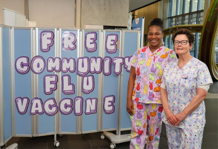Two female nurses wearing fun scrubs standing next to a promotional sign that reads "free community flu vaccine". 