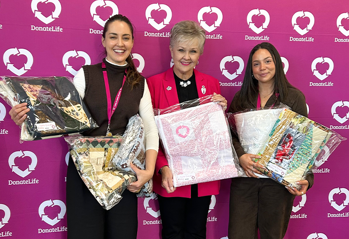 Leslie pictured in the middle, standing in front of a pink DonateLife media wall, holding some of the handmade quilts she is donating to DonateLife WA.