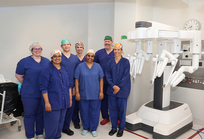 Seven theatre staff wearing blue theatre scrubs standing together next to the da Vinci robotic system.